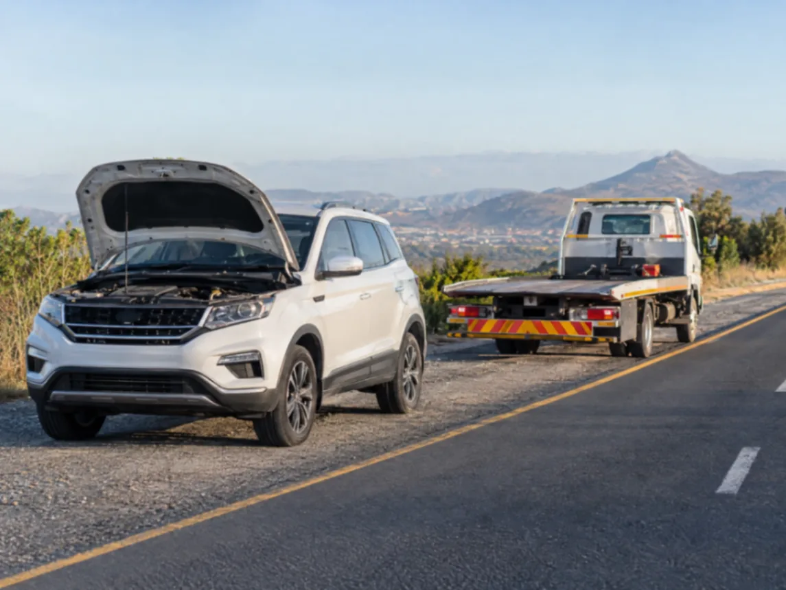White SUV with its bonnet open on the road shoulder, a flatbed tow truck parked ahead
